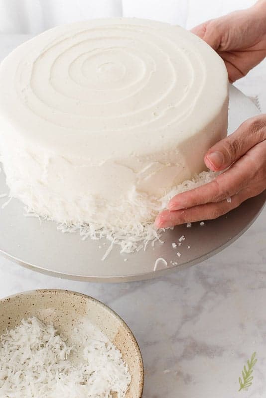 A hand presses coconut flakes against the side of the Piña Colada Cake