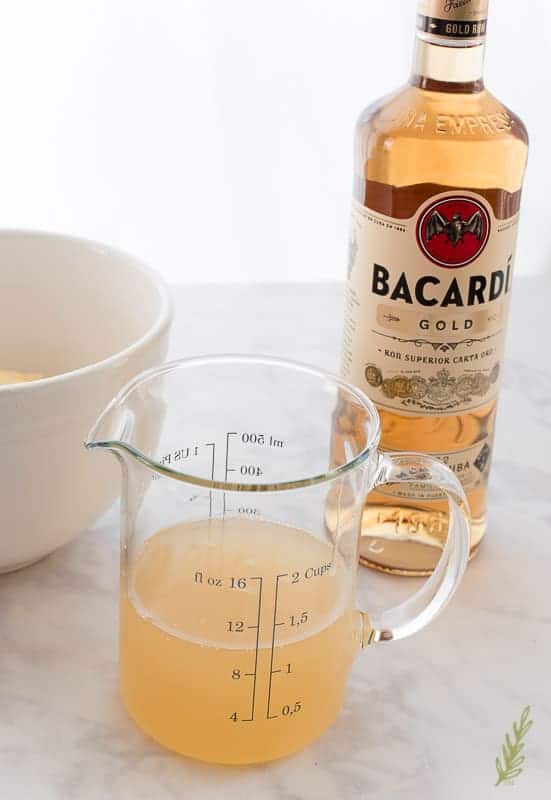 A clear, glass measuring cup filled with pineapple juice and rum sits in front of a bottle of rum which is in the background to the right. 