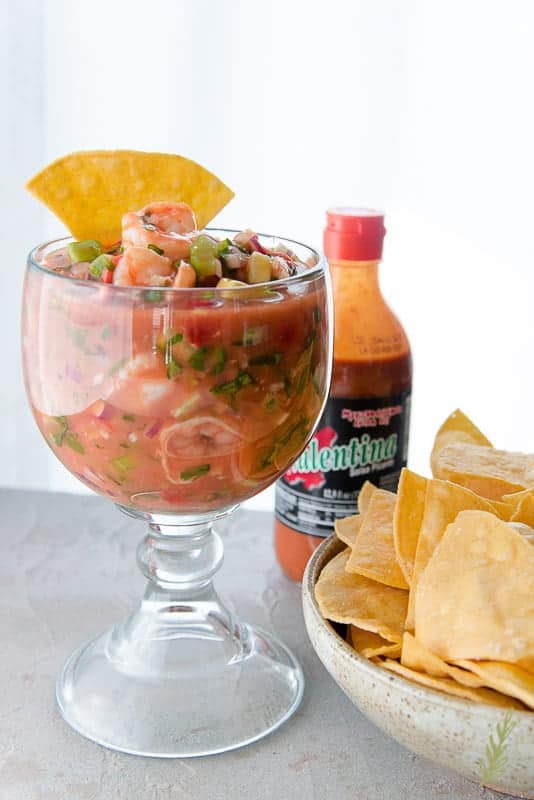 Goblet of Coctel de Camarones (Mexican Shrimp Cocktail) alongside a bowl of tortilla chips and a bottle of Valentina Mexican hot sauce.