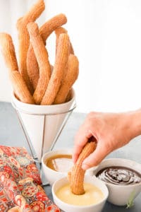 A hand dips a churro into a bowl of Crème Anglaise (Warm Vanilla Bean Sauce)