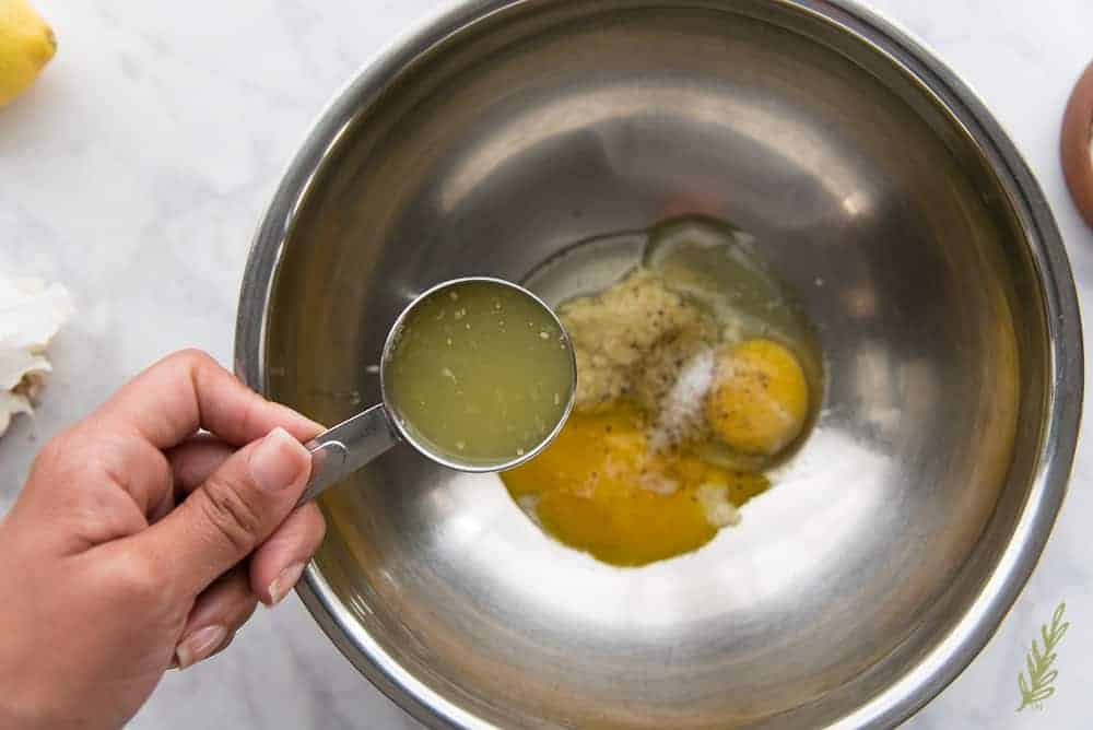 Adding lemon juice to the bowl with eggs, garlic, salt, pepper, and dijon mustard