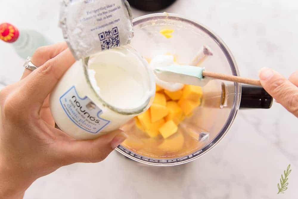 Adding the plain greek yogurt to the blender with the mango chunks.