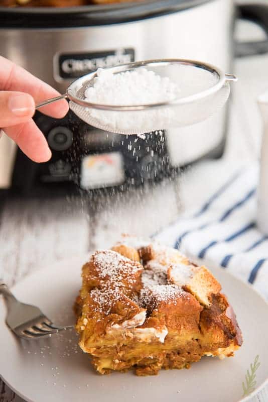 Sprinkling powdered sugar over a serving of Pumpkin Cream Cheese Slow Cooker French Toast Casserole, the slow cooker is in the background
