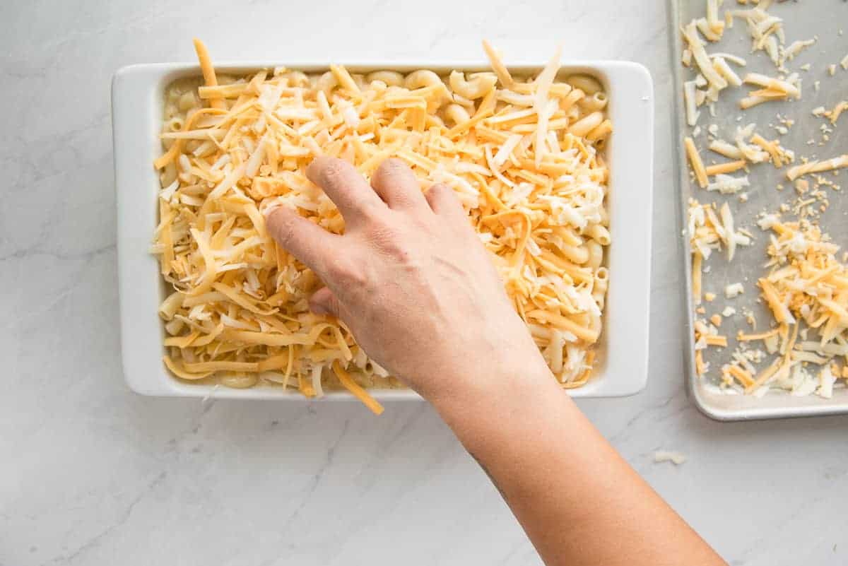 A hand sprinkles cheese over the panned 5 Cheese Baked Macaroni and Cheese in a baking dish.