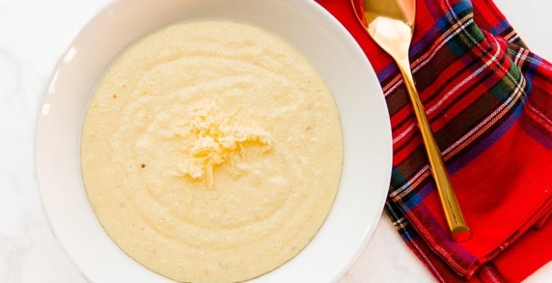 An overhead shot of a white bowl filled with Cheesy Grits with Garlic a red napkin lays next to it