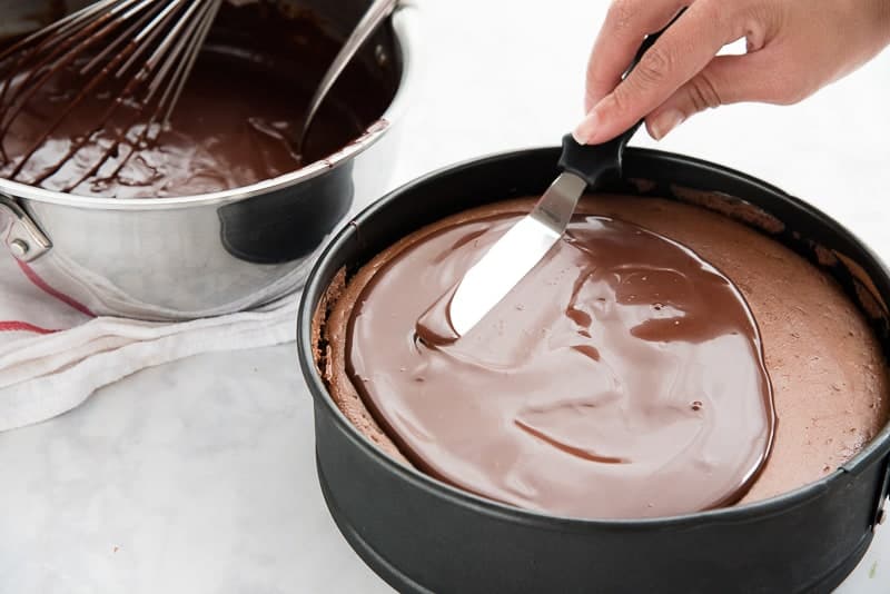 A hand uses a spatula to spread the chocolate ganache over the baked Nutella Cheesecake
