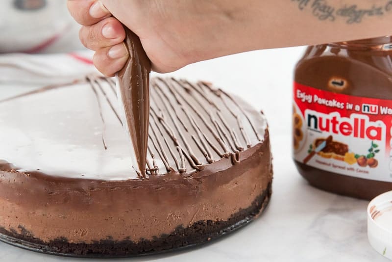 A hand drizzles Nutella onto the cheesecake