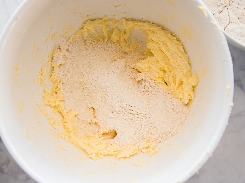 The dry ingredients are added gradually to the butter and sugar mixture in a white mixing bowl. 