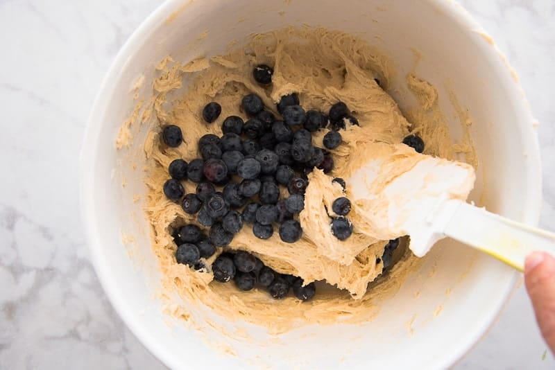 A white bowl filled with Blueberry-Almond muffin batter. A white spatula is stirring in blueberries