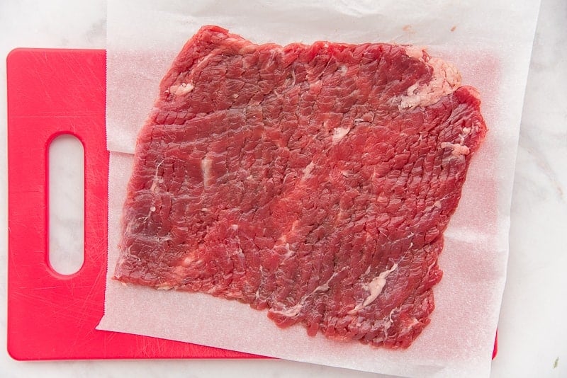The cube steak, after being flattened, sits on a red cutting board