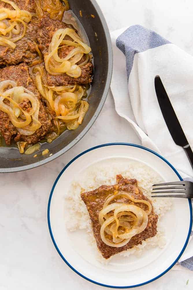 A plate with Bistec Encebollao and white rice sits next to a pan filled with bistec encebollao