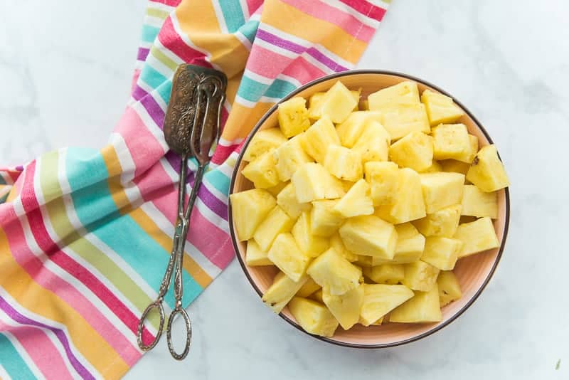 An overhead image of a pink bowl filled with pineapple chunks. A towel sits near the bowl underneath a pair of tongs