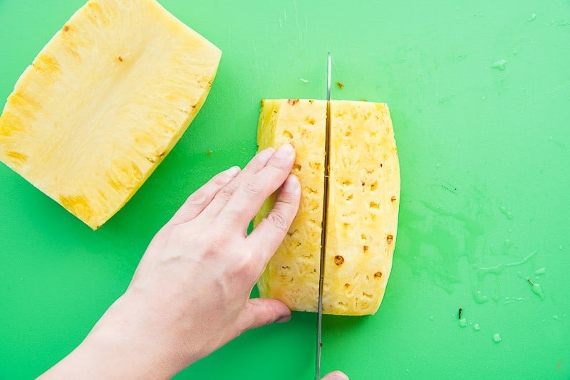 A hand is using a knife to cut the pineapple half into quarters on a green cutting board