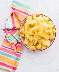 A portrait image of a pink bowl filled with cut pineapples. A multi-colored kitchen towel sits next to the bowl