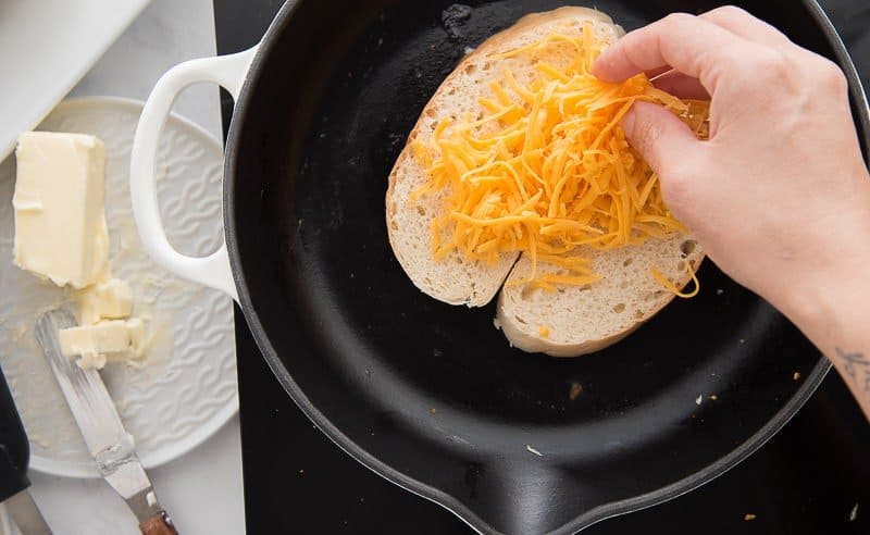 A hand sprinkle the shredded Queso de Papa onto the slices of bread in the pan