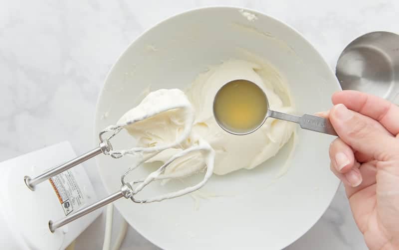 A hand holds a tablespoon of lemon juice over the white bowl containing the mixed cream cheese and sugar