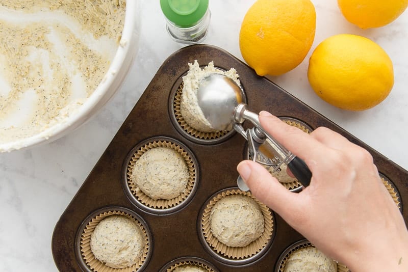 A hand uses a portion scoop to scoop the mixed batter into a muffin tin lined with paper cups