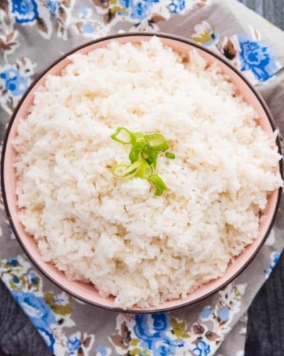 An overhead image of a bowl of Steamed White Rice on a grey floral napkin. A black spoon is next to the bowl
