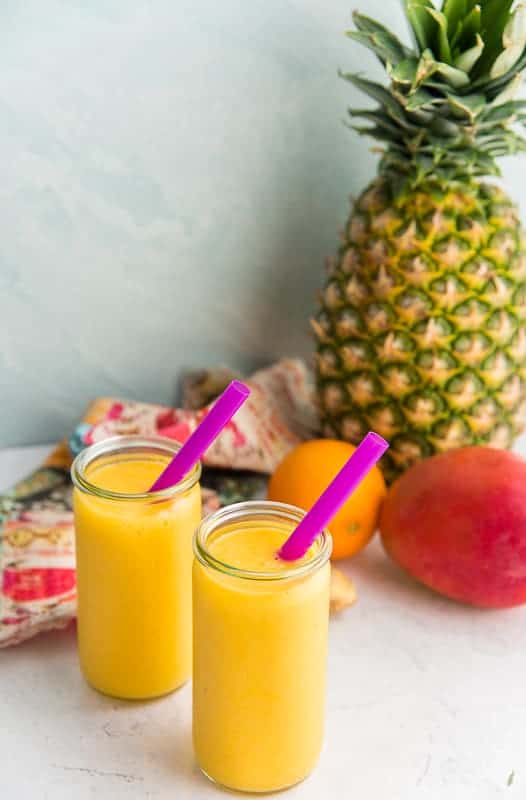 Two glasses filled with the vitamin C bomb smoothie. A pineapple, mango and orange in the background