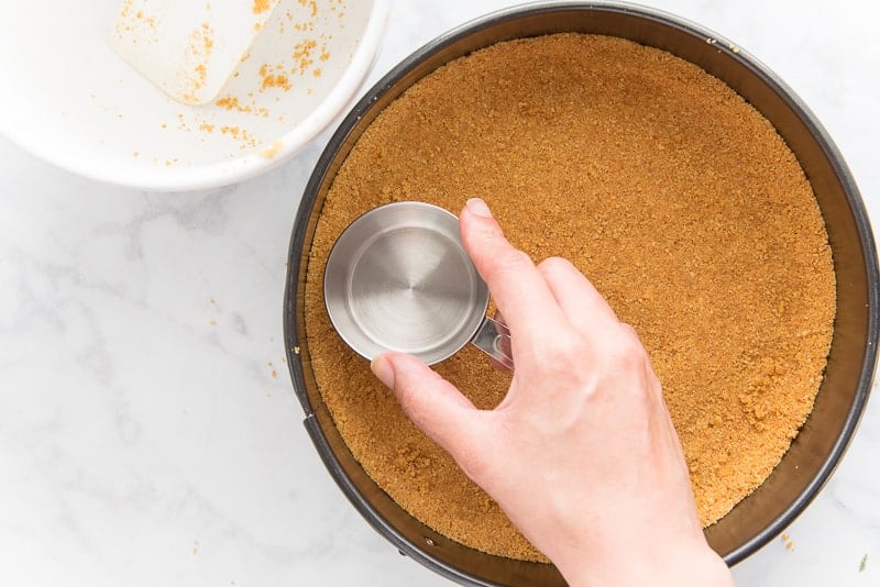 A measuring cup is used to press the graham cracker crumb mixture into the cheesecake pan