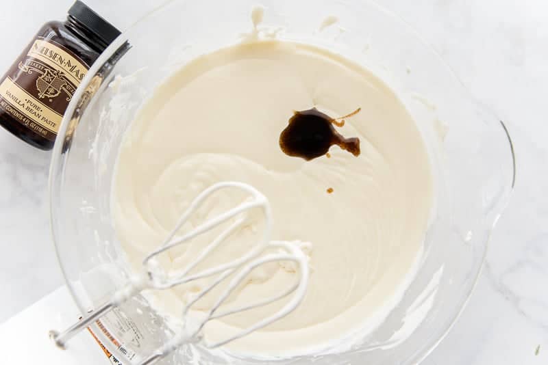 Vanilla bean paste is added to the bowl filled with batter, the bottle of vanilla bean paste is in the top left corner. The beaters of the mixer are seen in the bottom left corner. 