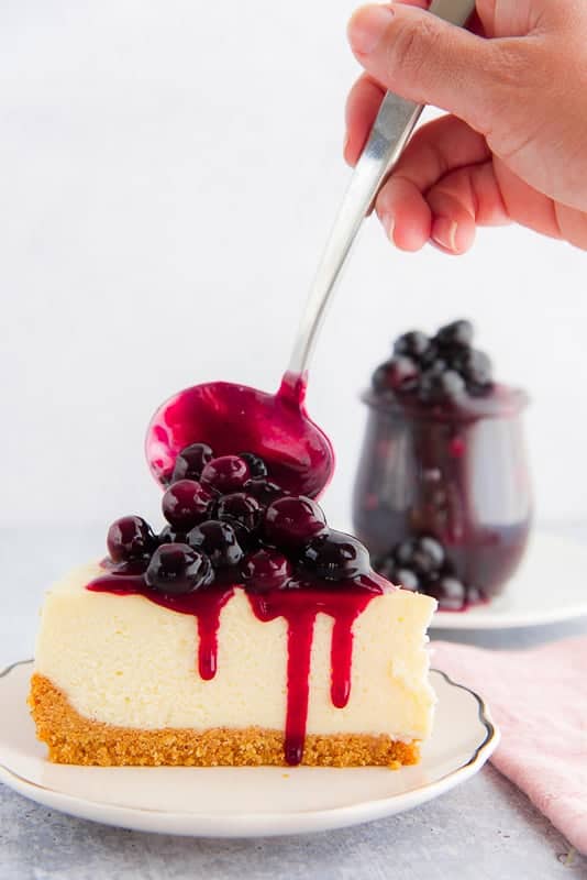 A jar of Blueberry Ginger Dessert Topping is behind a slice of cheesecake that is being topped with more topping