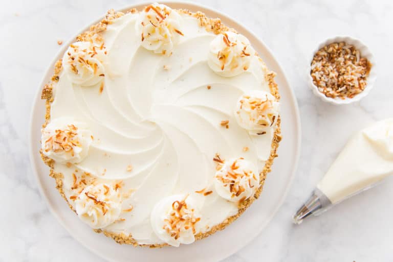 A top view of a coconut cream pie on a white plate, decorated with swirls of whipped cream and toasted coconut flakes. A small white bowl with more toasted coconut and a piping bag are beside the plate on a marble surface.