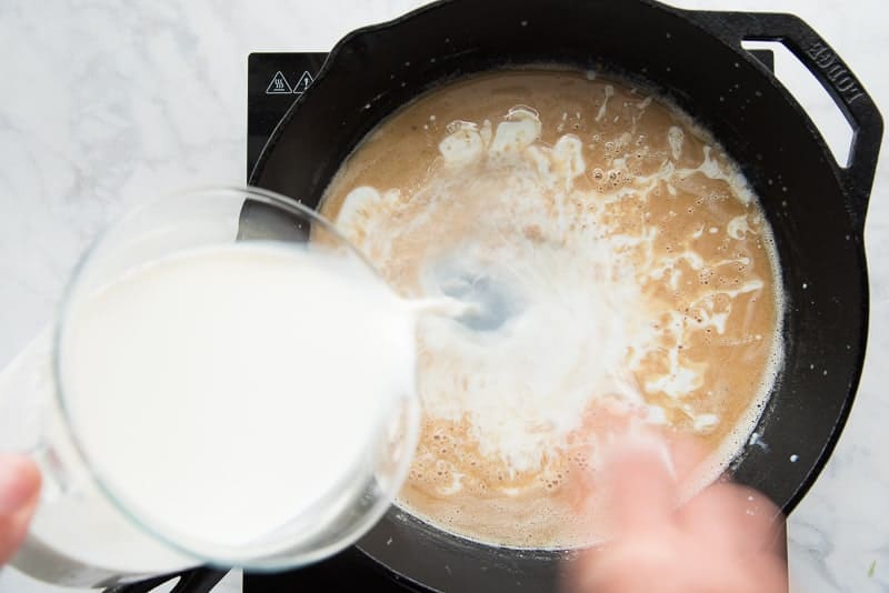 A pitcher of whole milk is poured into the brown roux in a cast iron skillet