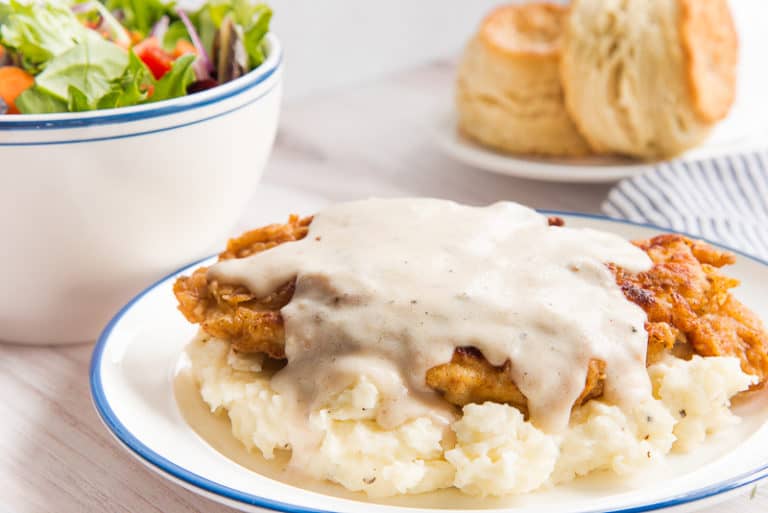 A plate of chicken fried steak topped with creamy gravy, served over mashed potatoes. In the background, there is a bowl of fresh salad with various vegetables and a plate with a biscuit. The meal is set on a white tabletop.