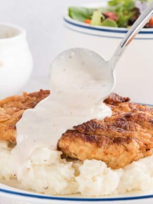 A hand ladles pan gravy over fried chicken and mashed potatoes