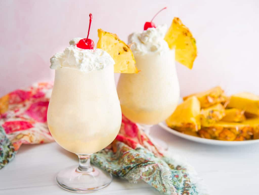 A horizontal image of two glasses filled with Piña Coladas next to a plate of sliced pineapples