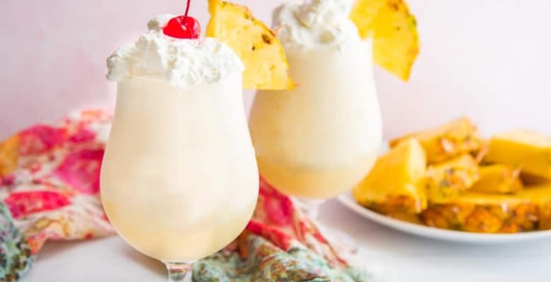 A horizontal image of two glasses filled with Piña Coladas next to a plate of sliced pineapples