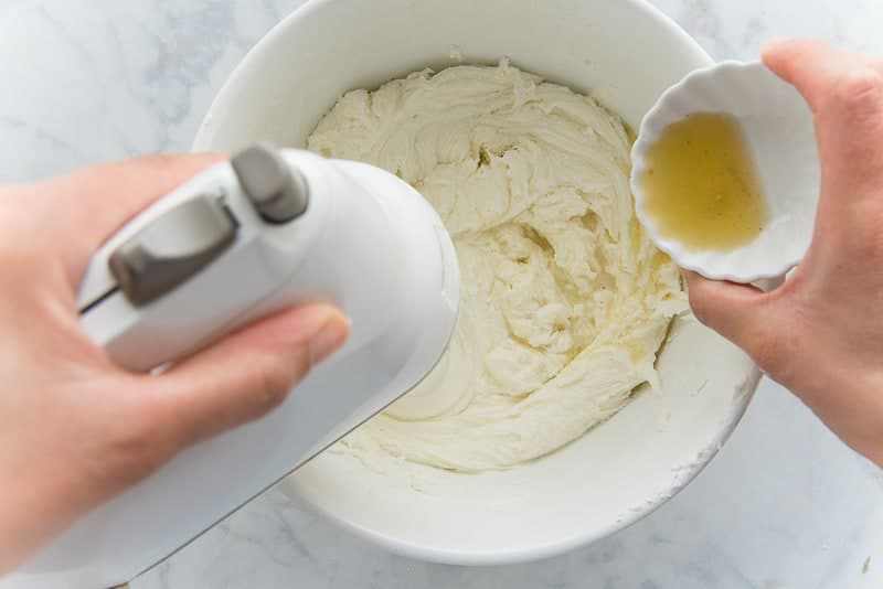 A hand adds lemon juice and vanilla extract to the mixing bowl