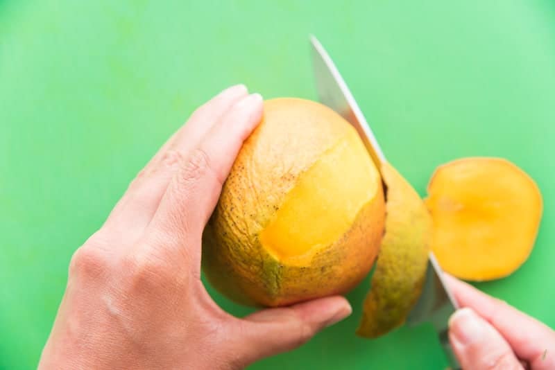 A hand holds a knife as the other holds the mango which is being peeled with the knife