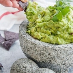 A hand uses a blue corn tortilla chip to scoop some guacamole
