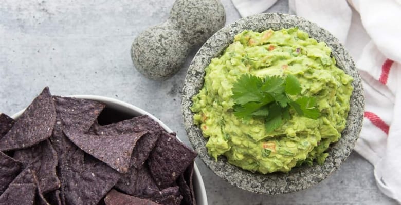A horizontal image of a bowl of guacamole next to a bowlful of chips