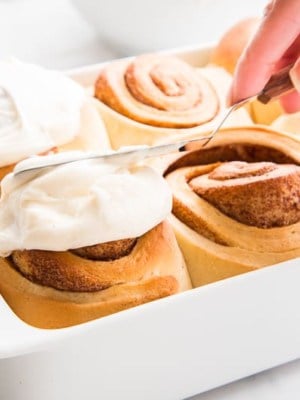 A hand spreads Cream Cheese Icing over a Cinnamon Roll that's in a white casserole dish