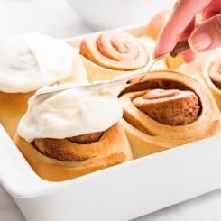 A hand uses a spatula to spread Cream Cheese Icing on the Cinnamon Rolls in a white dish