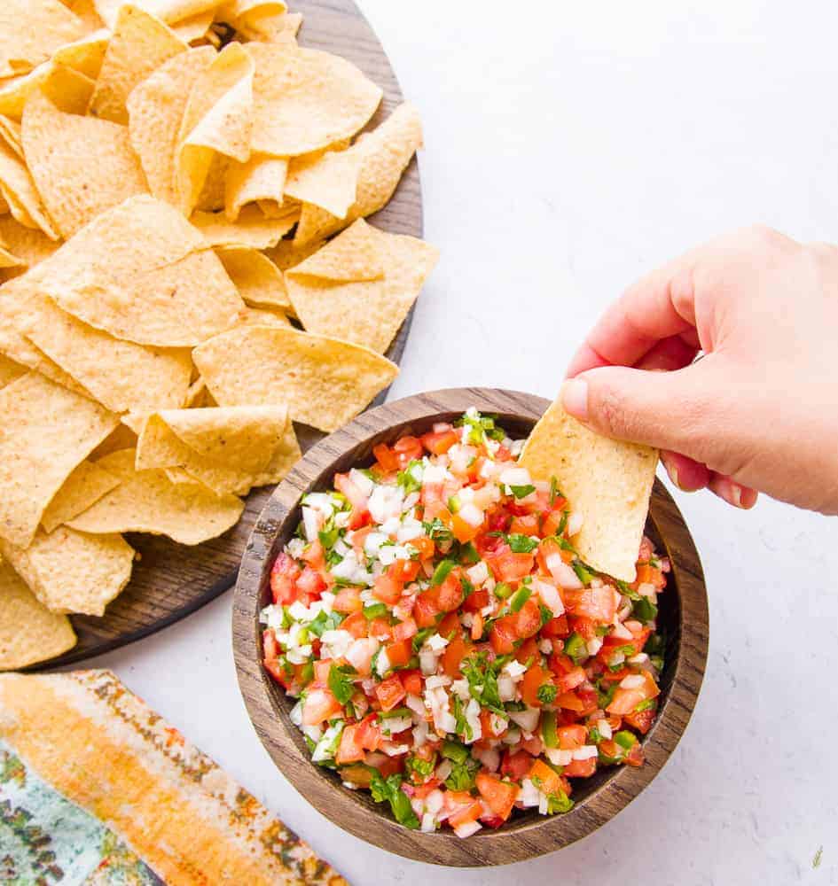 A hand uses a corn tortilla chip to scoop pico de gallo from a wooden bowl