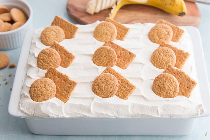 A white dish filled with Homemade Banana Pudding, topped with whipped cream, graham cracker pieces and vanilla wafers. A half-peeled banana is partially sliced sitting on a wooden board at the top of the image. A white bowl filled with vanilla wafers sits in top left corner