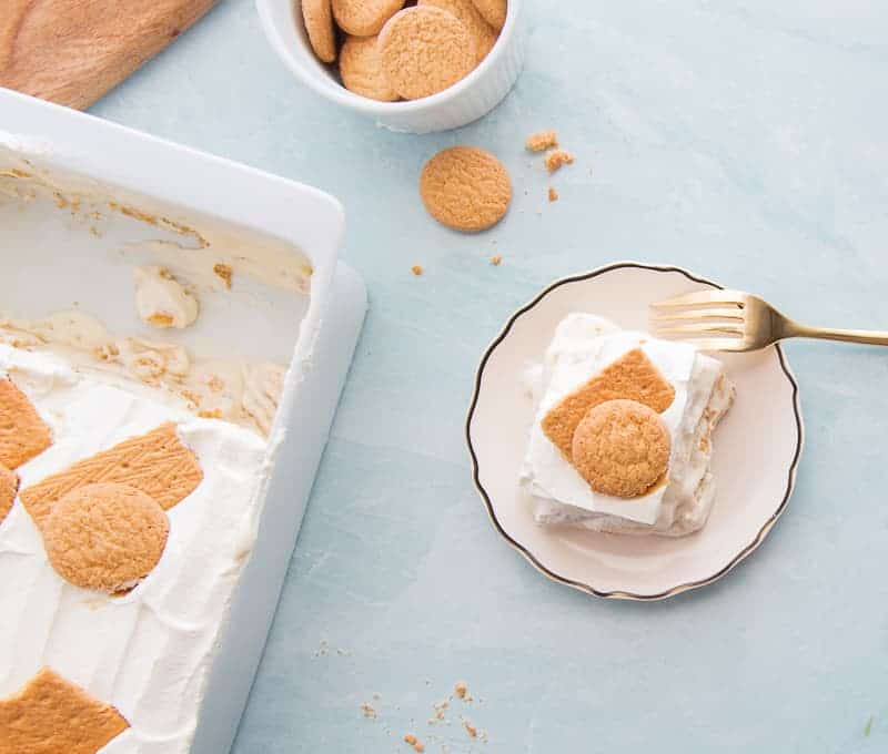 An overhead image of a white, gold-rimmed plate with a portion of Homemade Banana Pudding on it. A gold fork rests on the plate. A white casserole dish with banana pudding portions removed is in the bottom left corner. A white bowl with vanilla wafers is in the top left corner