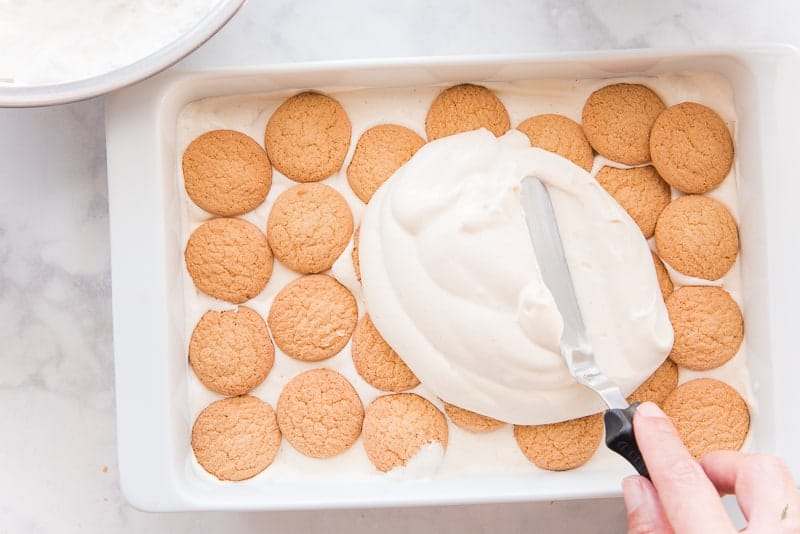 Pastry cream is being spread over a layer of vanilla wafers which is in a white casserole dish a metal bowl with whipped cream sits in top left corner