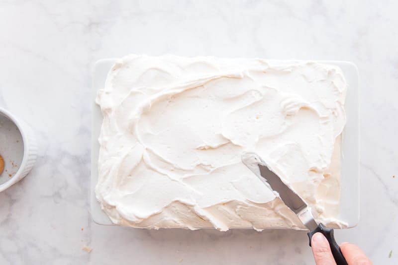 A hand uses a metal spatula to smooth whipped cream on to the pudding, which is in a white baking dish.