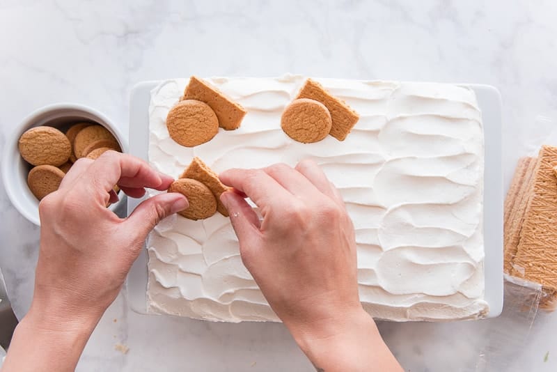 Two hands arrange a piece of a graham cracker and a vanilla wafer to garnish the whipped cream topped Homemade Banana Pudding. It is in a white baking dish. A white bowl of vanilla wafers is to the left of the baking dish, a packet of graham crackers is on the right
