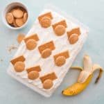 An overhead image of the Homemade Banana Pudding. In a white ceramic baking dish and garnished with graham cracker pieces and vanilla wafers. A partially peeled banana sits at bottom right. A white bowl filled with vanilla wafers is in the top left corner.