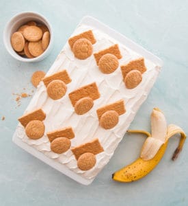 An overhead image of the Homemade Banana Pudding. In a white ceramic baking dish and garnished with graham cracker pieces and vanilla wafers. A partially peeled banana sits at bottom right. A white bowl filled with vanilla wafers is in the top left corner.
