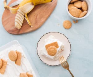 An overhead image of a portion of Homemade Banana Pudding on a white, gold-rimmed plate. A sliced, half peeled banana is on a wooden cutting board in top left. A white bowl filled with vanilla wafer in top right corner. A corner of the pan filled with banana pudding in bottom left. A gold spoon rests on the plate