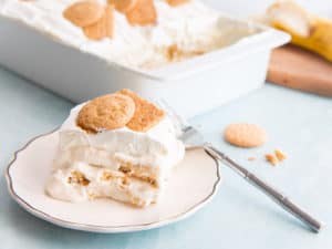 A serving of Homemade Banana Pudding on a white, gold-rimmed plate. A silver fork is propped on the plate. A white casserole dish filled with banana pudding and a wooden cutting board with a sliced banana is in the background