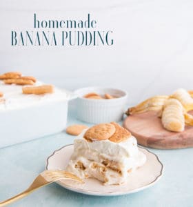 A long pin image of a white, gold-rimmed plate with a portion of Homemade Banana Pudding on it. Gold fork rests on plate. In right background a sliced banana on a wooden board, middle background a white bowl of vanilla wafers, left background a white dish filled with the Homemade Banana Pudding. Blue text that reads "Homemade Banana Pudding" is in top left corner
