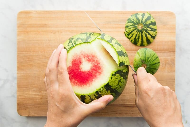 The rind of a watermelon is cut away from a whole melon on a wooden cutting board